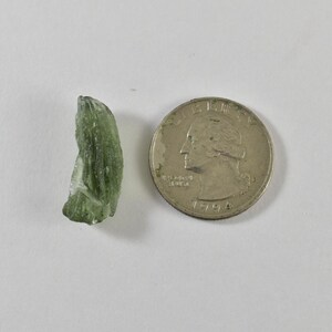 May include: A small, translucent, olive-green moldavite stone with a textured, irregular surface next to a silver-coloured United States quarter coin dated 1994, which provides a clear visual scale for the size of the raw mineral specimen.