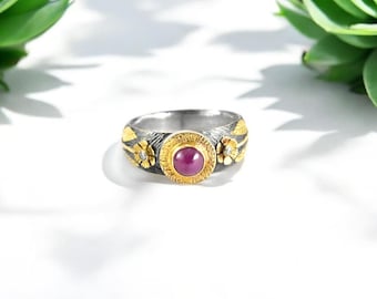 May include: A silver ring with gold accents and a central, round, red gemstone. The ring features floral and leaf designs, with small clear stones set into the flowers. The ring is set against a white background.
