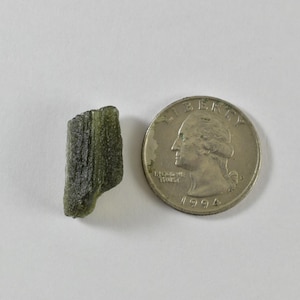 May include: A small, rough, olive-green moldavite fragment with a textured, irregular surface positioned next to a silver-coloured United States quarter coin for scale, showing the relative size of the mineral specimen against the currency.