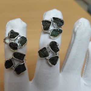 Peut inclure: Quatre bagues en argent, chacune ornée d'un groupe de pierres vertes foncées de forme irrégulière. Les bagues sont présentées sur une surface blanche, mettant en valeur le contraste entre l'argent et les pierres.