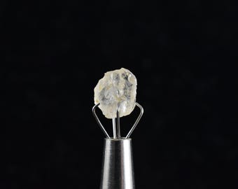 May include: A clear, faceted, rough-cut diamond held in a metal clamp against a black background.