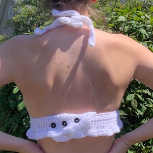 May include: A white, handmade crochet halter top. The top features a halter neck tie and a ruffled bottom edge. Three dark buttons secure the back closure. The garment is made of white yarn.