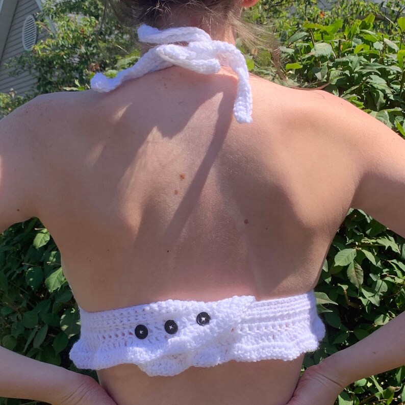 May include: A white, handmade crochet halter top. The top features a halter neck tie and a ruffled bottom edge. Three dark buttons secure the back closure. The garment is made of white yarn.