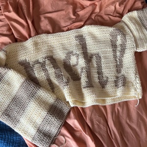 May include: A crocheted blanket with a striped pattern in beige and white. The blanket has the word "LOVE" crocheted into it in a darker beige color.