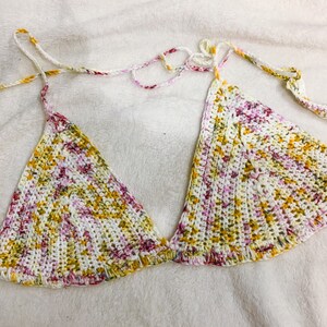 May include: A handmade crochet bikini top in a triangle style. The top features a colorful pattern with white, yellow, and pink threads. The straps are white with accents of yellow, pink, and red. The garment is designed for swimwear.