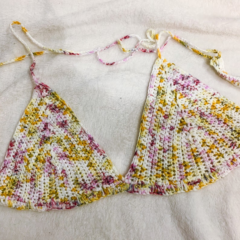May include: A handmade crochet bikini top in a triangle style. The top features a colorful pattern with white, yellow, and pink threads. The straps are white with accents of yellow, pink, and red. The garment is designed for swimwear.