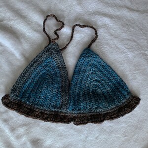 May include: A handmade crochet top in shades of blue and gray, with a brown ruffled trim and straps. The triangle-shaped cups are connected by brown straps that tie at the neck. The top is made of soft yarn.