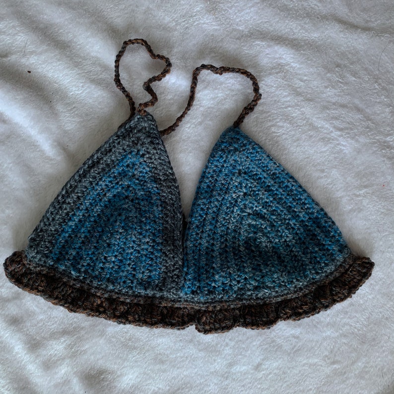 May include: A handmade crochet top in shades of blue and gray, with a brown ruffled trim and straps. The triangle-shaped cups are connected by brown straps that tie at the neck. The top is made of soft yarn.