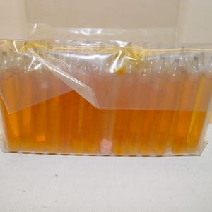 May include: A rectangular clear plastic package containing a row of upright, slender, cylindrical glass ampoules filled with a translucent amber-colored liquid. One ampoule in the center contains a small, light pink solid object at the base.