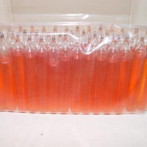 May include: A clear plastic bag containing a large quantity of small, cylindrical glass ampoules filled with a vibrant orange liquid. Each ampoule features a tapered, sealed top and a flat base, arranged in a dense, upright formation within the packaging.