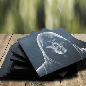 May include: A set of black slate coasters with a white drawing of a hooded figure with a skull face. The coasters are stacked on a wooden surface.