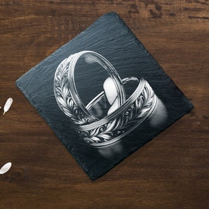 May include: Two silver wedding bands with intricate leaf designs are displayed on a black slate coaster. The bands are intertwined and the design is detailed.
