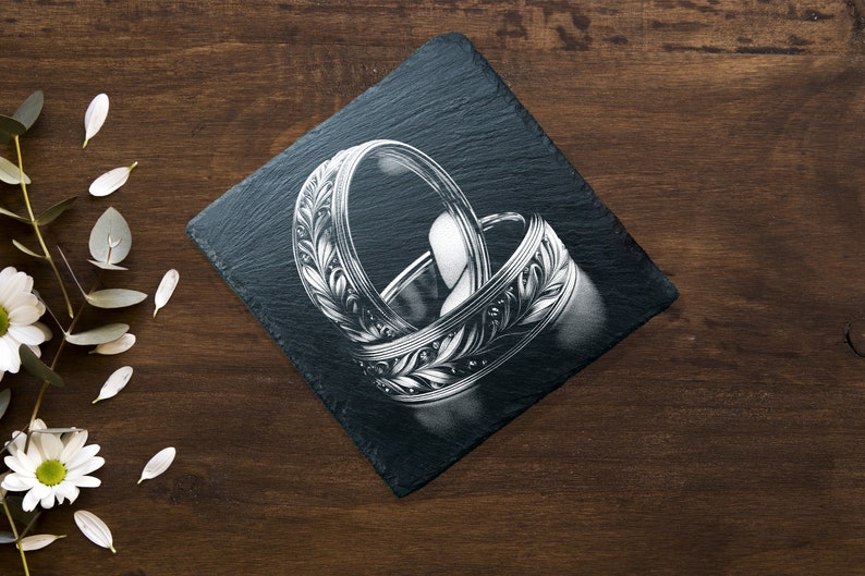 May include: Two silver wedding bands with intricate leaf designs are displayed on a black slate coaster. The bands are intertwined and the design is detailed.