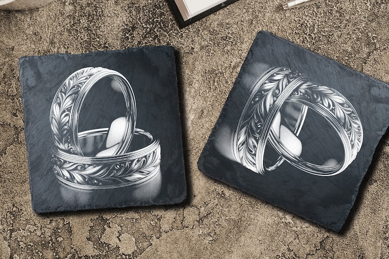 May include: Two silver rings with intricate floral designs are displayed on black slate coasters. The rings are stacked on top of each other, showcasing the detailed engravings.