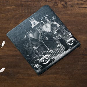 May include: A black and white image of a spooky house with a witch and pumpkins. The house is decorated with cobwebs and bats. The image is printed on a black slate coaster.