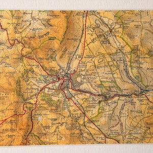 May include: A framed vintage map of Buxton, Derbyshire, showing detailed topography and red road markings. The map has a sepia-toned colour palette and is set within a white frame, highlighting the town's layout and surrounding areas.