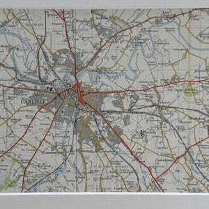 May include: A framed map of Carlisle, Cumbria, showing the city and its surroundings. The map is in beige, grey and red, with roads and geographical features clearly marked. The map is encased in a white frame, suitable for home decor.
