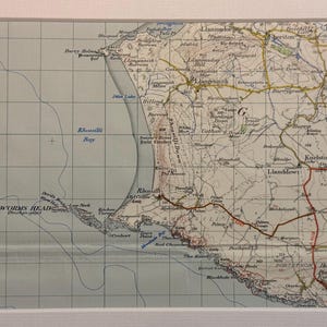 May include: A vintage map of the Gower Peninsula in Wales, UK. The map shows the coastline, towns, and roads. The map is printed on paper and has a light blue background with brown lines and text.