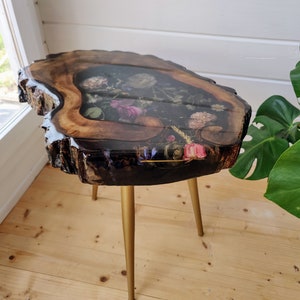 Può includere: Un tavolo rotondo in legno con un bordo vivo e gambe in metallo dorato. Il piano del tavolo è in resina e presenta fiori e foglie pressati.