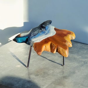 Op de afbeelding: Een kleine, onregelmatig gevormde houten tafel met een blauw, wit en zwart harsblad. De tafel wordt ondersteund door drie dunne metalen poten.