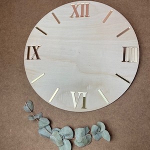 Könnte beinhalten: Ein leeres Holz-Zifferblatt mit römischen Ziffern in Gold. Das Zifferblatt befindet sich auf einem braunen Hintergrund mit einem Zweig grüner Eukalyptusblätter.