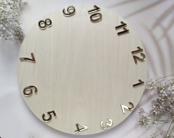 Peut inclure: Un cadran d'horloge rond en bois non fini avec des chiffres métalliques dorés. Les chiffres de 1 à 12 sont espacés uniformément autour du périmètre du cadran. Le bois est de couleur claire et naturelle.