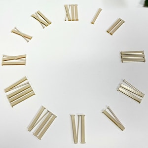 Può includere: Un set di numeri romani dorati per orologio, disposti in cerchio su uno sfondo bianco.