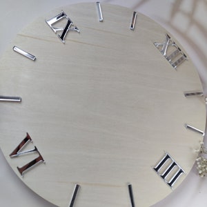 Op de afbeelding: Een wit houten klok gezicht met zilveren Romeinse cijfers als uurmarkeringen. Het klok gezicht is leeg, zonder wijzers of andere markeringen.