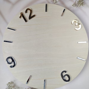 Könnte beinhalten: Ein leeres Holzuhrblatt mit silbernen und goldenen Zahlen. Die Zahlen 12, 3, 6 und 9 sind sichtbar. Das Uhrblatt ist hellbraun und hat eine glatte Oberfläche.