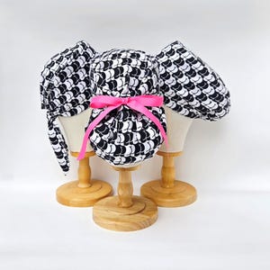 May include: Three scrub caps featuring a repeating black and white pattern of stylized buttocks. The center cap is gathered at the back and secured with a bright pink ribbon tied in a bow. Each cap is displayed on a wooden mannequin head stand.