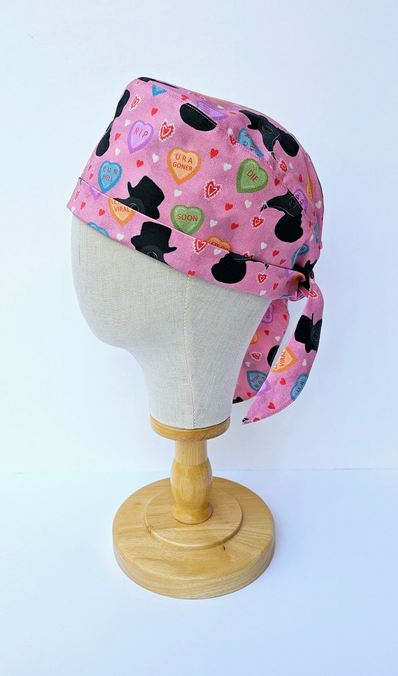May include: Pink surgical scrub hat with a black and white print of hearts, hats, and the words "RIP", "Gun Hell", "Viral", "URA Goner", "Soon", and "Die".