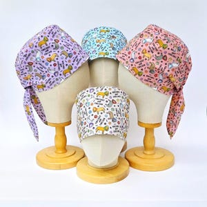 May include: Four patterned surgical scrub caps on mannequin heads. The caps feature dogs, paw prints, and the text "Bitches get stitches". Colours include lavender, light blue, pink, and white. The caps are displayed on wooden stands.