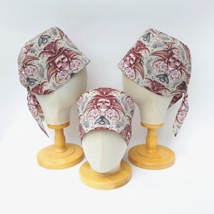 May include: Three fabric head coverings featuring a repeating pattern of skulls wearing plague doctor masks, roses, beetles, and centipedes in shades of red, black, and grey on a light beige background. Each cap is displayed on a mannequin head.