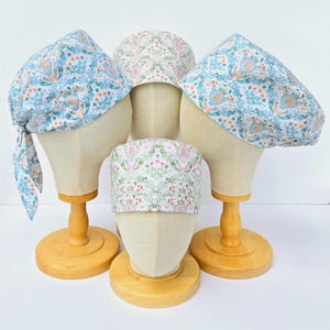 以下が含まれることがあります： 青、緑、ピンク、白の色の花柄が施された手術帽が4つあります。帽子は木製の人形に飾られています。