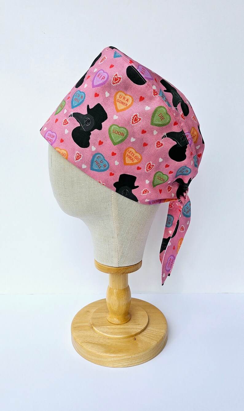 May include: Pink surgical scrub hat with a black and white print of a plague doctor and candy hearts with phrases like "RIP", "URA GONER", "SOON", "C U IN HELL", "DROP DEAD", "LETS GO VIRAL", and "DIE".