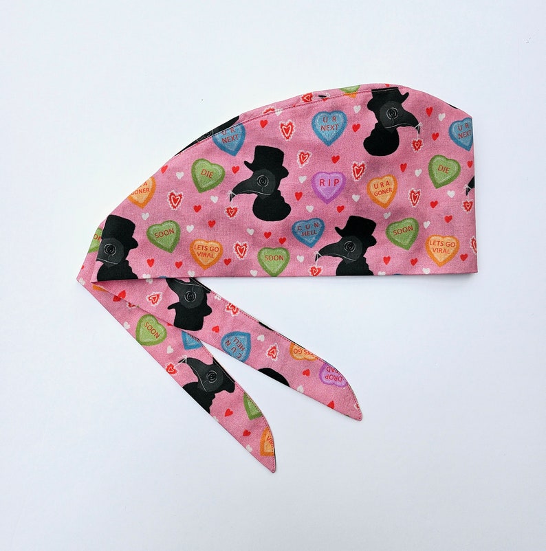 May include: Pink surgical scrub hat with a repeating pattern of black plague doctor masks and candy hearts with phrases like "RIP", "Let's Go Viral", and "U R Next".