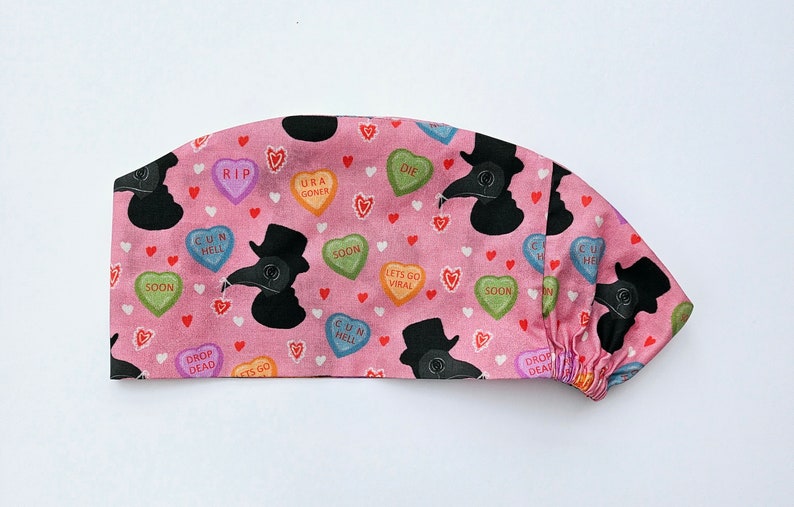 May include: Pink surgical scrub hat with a pattern of black plague doctor silhouettes and candy hearts with phrases like "RIP", "URA GONER", "CUR HELL", "SOON", "LETS GO VIRAL", "DROP DEAD", and "CUN HELL".
