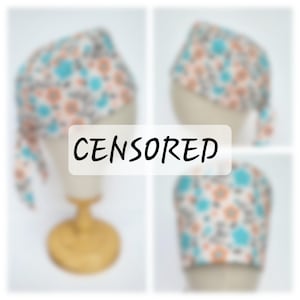 Op de afbeelding: Een witte stoffen hoofdband met een bloemenpatroon in tinten blauw, oranje en grijs. De band is aan de achterkant van het hoofd gebonden en heeft een lange staart die naar beneden hangt.