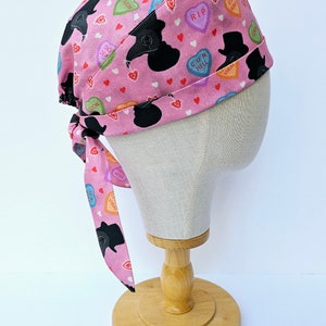 May include: Pink surgical scrub hat with a tie, featuring a pattern of black silhouettes and colorful candy hearts with phrases like "RIP", "CUN HELL", and "SOON".