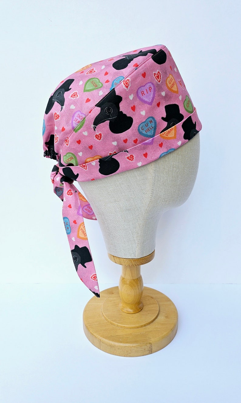 May include: Pink surgical scrub hat with a tie, featuring a pattern of black silhouettes and colorful candy hearts with phrases like "RIP", "CUN HELL", and "SOON".