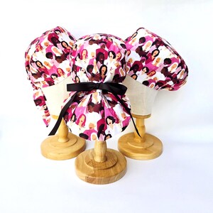 May include: Three scrub hats with a pink and white pattern, each with a black bow, displayed on wooden stands. The fabric design features illustrations of women with pink ribbons, representing breast cancer awareness. Designed for medical use.