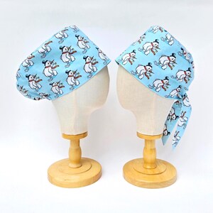 May include: Two light blue surgical scrub caps with a repeating pattern of snowmen wearing top hats and red scarves. The caps are displayed on mannequin heads with wooden stands. One cap is tied in a bow.