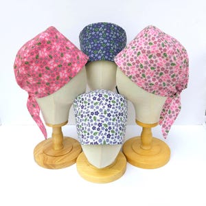 May include: Four patterned head coverings on wooden stands. Two are pink with floral designs, one is navy blue with a floral pattern, and one is white with blue and purple flowers. The head coverings are tied at the back.