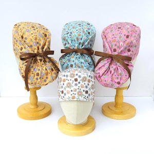 May include: Three surgical caps with a floral print featuring a repeated pattern of a buttock. The caps are tied with brown ribbon and are displayed on wooden stands.