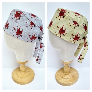 Könnte beinhalten: Zwei OP-Hauben, eine graue und eine beige, jeweils mit einem Muster aus roten Blutspritzern verziert. Die Hauben werden auf Schaufensterpuppenköpfen präsentiert, um ihr Design hervorzuheben. Die Bindebänder sind sichtbar.