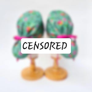 Può includere: Due cuffie chirurgiche con un motivo floreale verde acqua e accenti rosa sono esposte. Le cuffie sono legate con nastri rosa e poggiano su supporti di legno. La parola "CENSORED" è scritta sull'immagine in un rettangolo bianco.