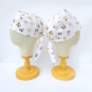 May include: Two white fabric headbands with a pattern of colourful people in various poses. The headbands are tied at the back and are displayed on two wooden mannequins.