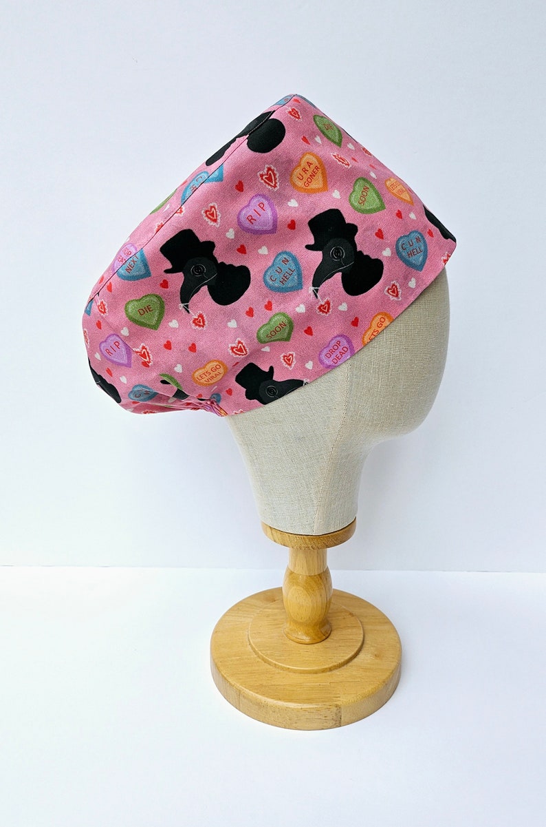 May include: Pink surgical scrub hat with a pattern of black silhouettes of people wearing top hats and holding a bird's beak mask. The hearts have various sayings on them, including "RIP", "DIE", "CUN HELL", "URA GONER", "SOON", "DROP DEAD", and "LETS GO VIRUNK".