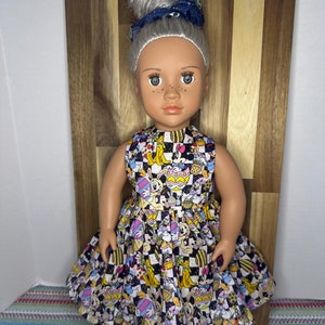 May include: A doll wearing a colorful dress with a checkered pattern and cartoon characters. The dress features Mickey Mouse, Minnie Mouse, Donald Duck, Goofy, and Pluto. The doll has gray hair and blue eyes.