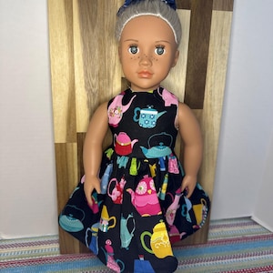 May include: A doll with gray hair styled in a bun, wearing a black dress patterned with colorful teapots. The doll has blue eyes and freckles. A blue bow is in the doll's hair. The doll is standing in front of a wooden background.